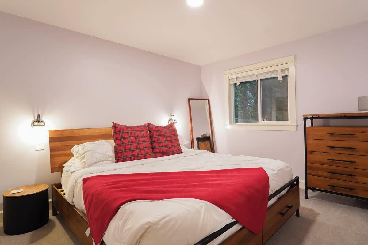Un dormitorio con cama de madera, colcha roja y blanca, cómoda de madera, mesita de noche redonda y una ventana con persianas.