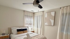 Et soveværelse med seng, ventilator, natbord, lampe, et vindue med gardiner og et tæppe.