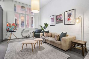 A modern living room with a sofa, two small tables, a floor lamp, and a large window with a view of a brick building. - Chic 2-Bed Apartment in Melbourne CBD (Melbourne)