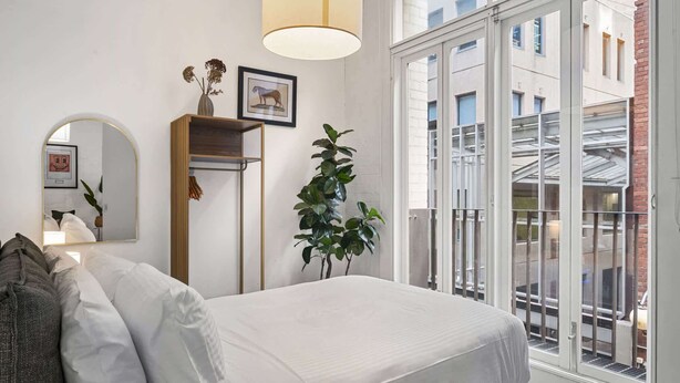 Quarto com cama, espelho, planta e janela com vista para edifícios.