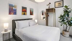 Quarto de hotel com cama, mesa de cabeceira, luminária, espelho, guarda-roupa e obras de arte nas paredes.