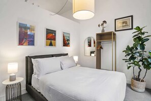 A hotel room with a bed, a nightstand, a lamp, a mirror, a wardrobe, and wall art. - Chic 2-Bed Apartment in Melbourne CBD (Melbourne)