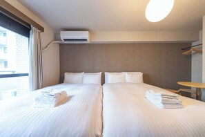 Quarto de hotel com duas camas, uma janela com cortinas, aparelho de ar condicionado e uma mesa com cadeira.