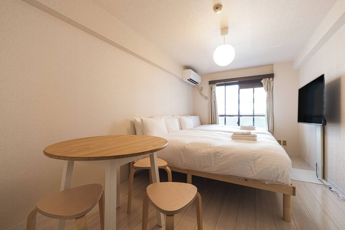 A bedroom with a bed, a round wooden table, two wooden chairs, a TV mounted on the wall, and an air conditioning unit.