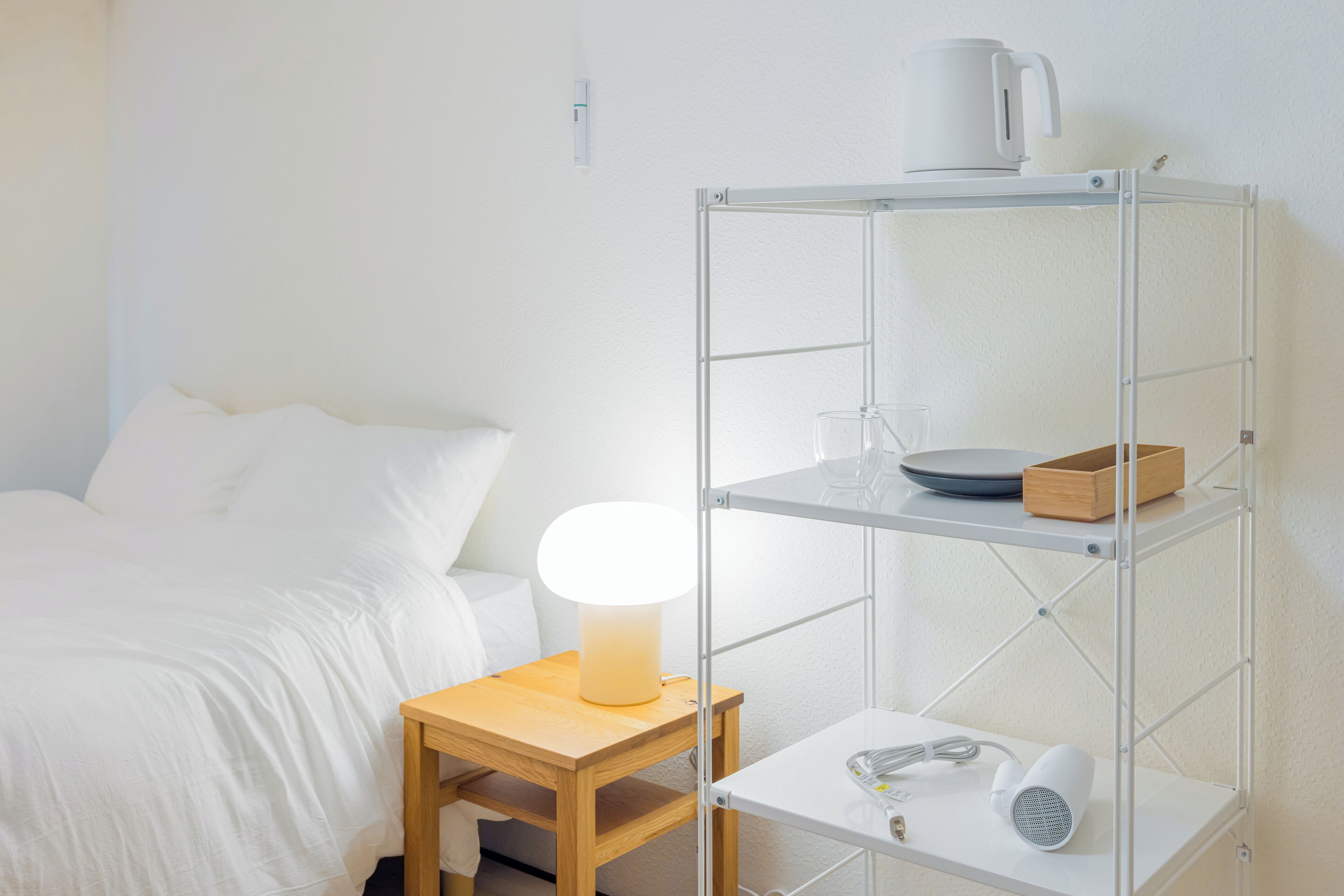 A hotel room with a bed, a bedside table with a lamp, a white shelf with a kettle, a box, and a hairdryer.