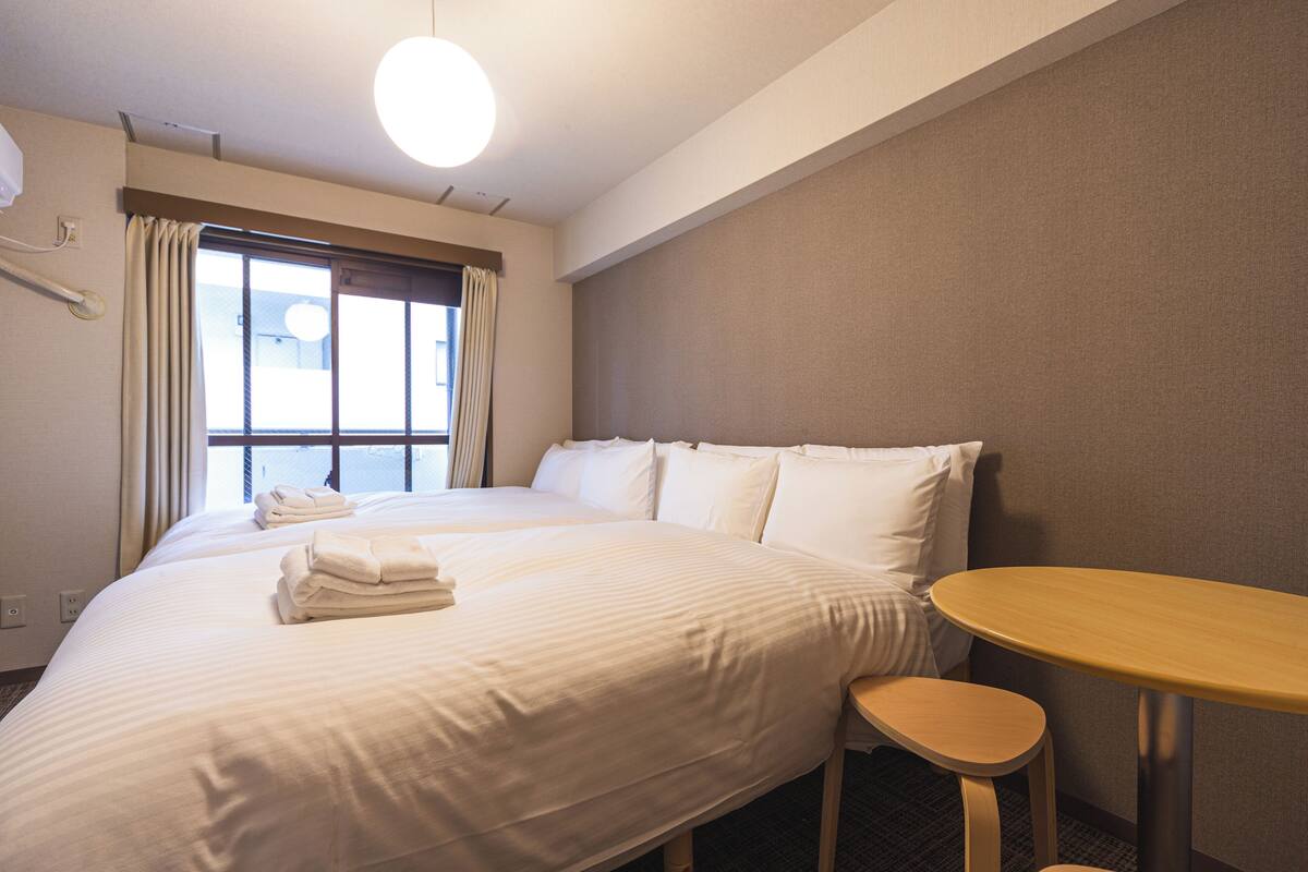 A hotel room with two beds, a small wooden table, and a window with curtains.