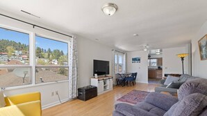 Een moderne woonkamer met een groot raam, een flatscreen-tv, een bank, een eethoek en een keuken op de achtergrond.