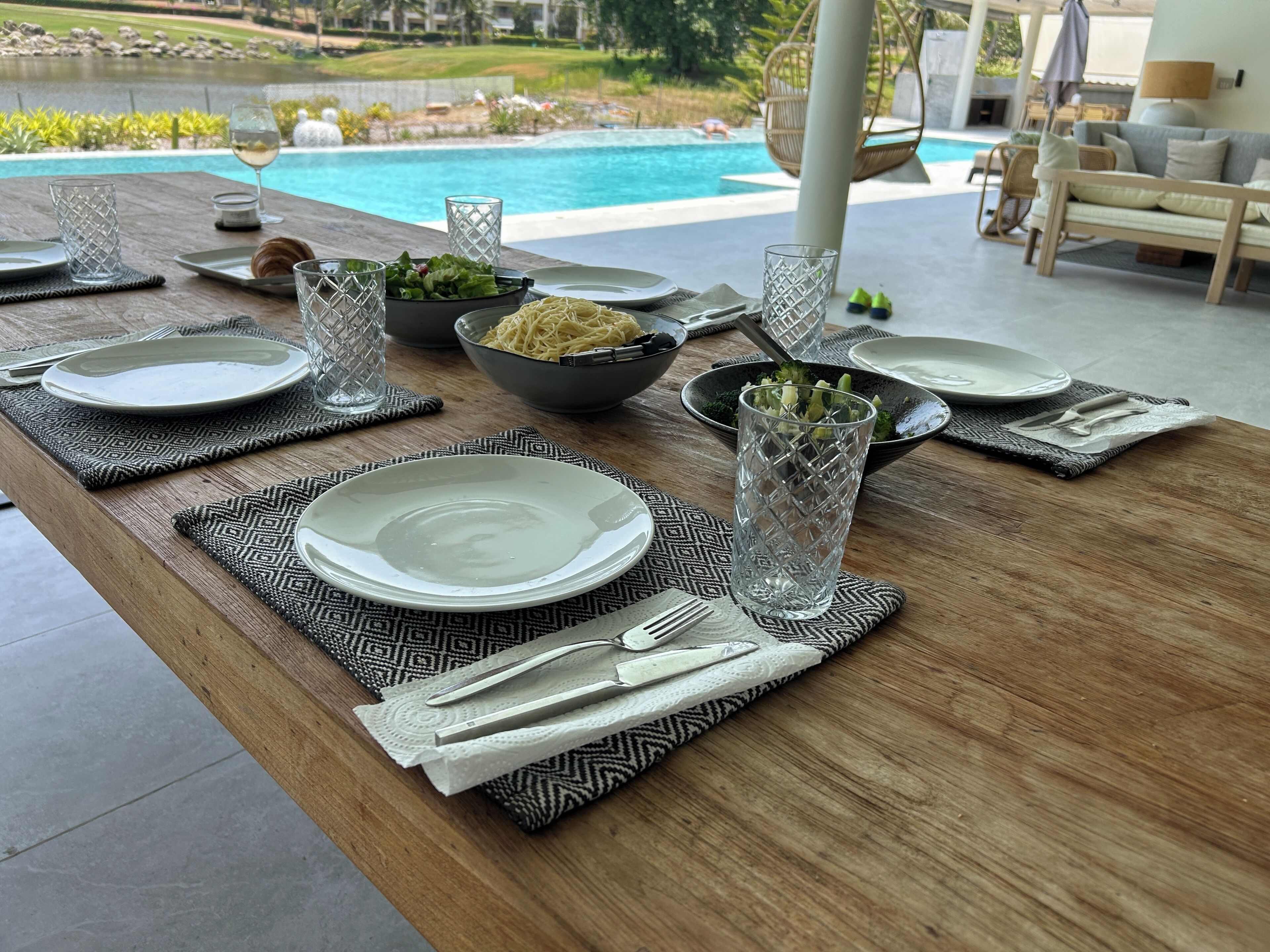 Mesa de comedor lista con platos, vasos y cubiertos, con una piscina y zona de estar exterior al fondo.