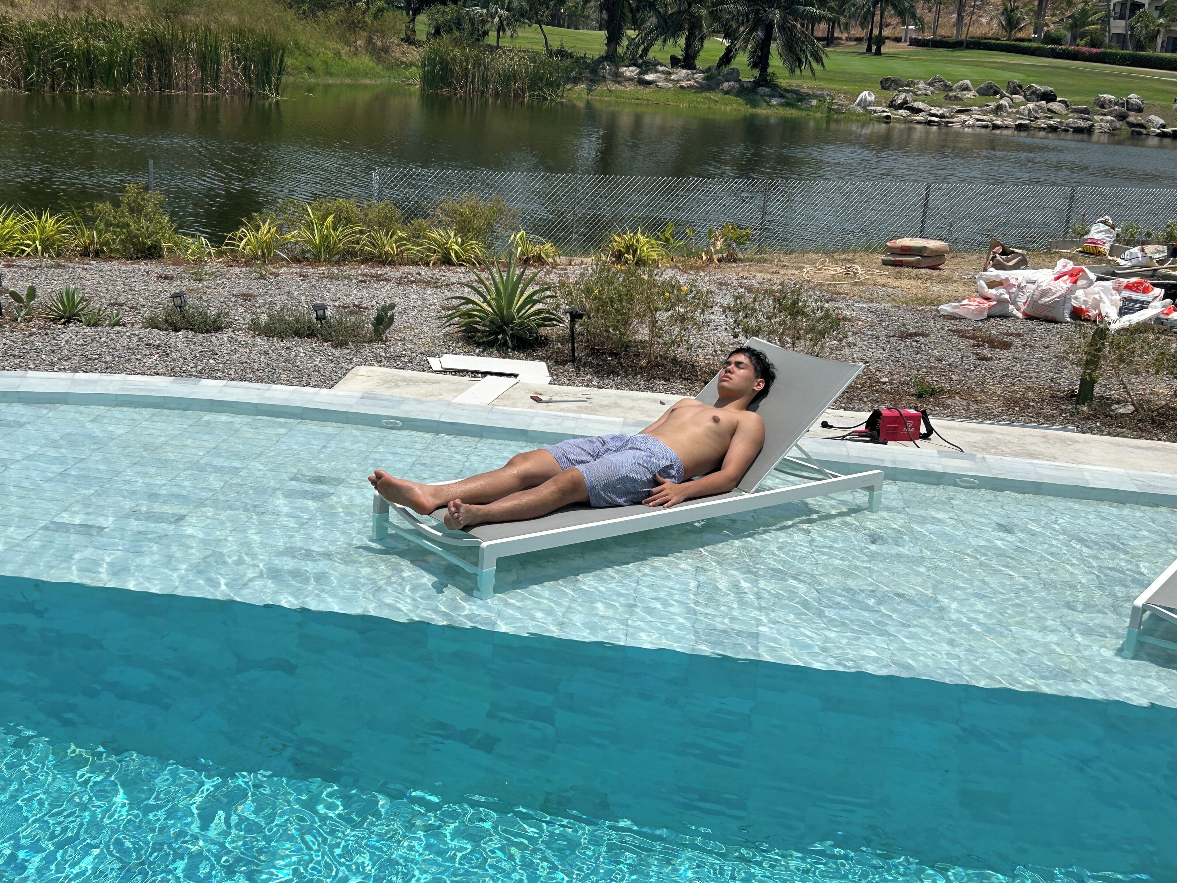 A pool with clear water, a person lying on a lounge chair, and a pond in the background.