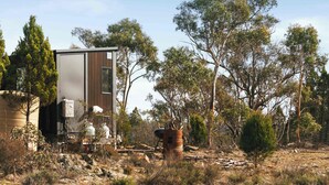 A rustic cabin with a large window, surrounded by trees and a rocky landscape.