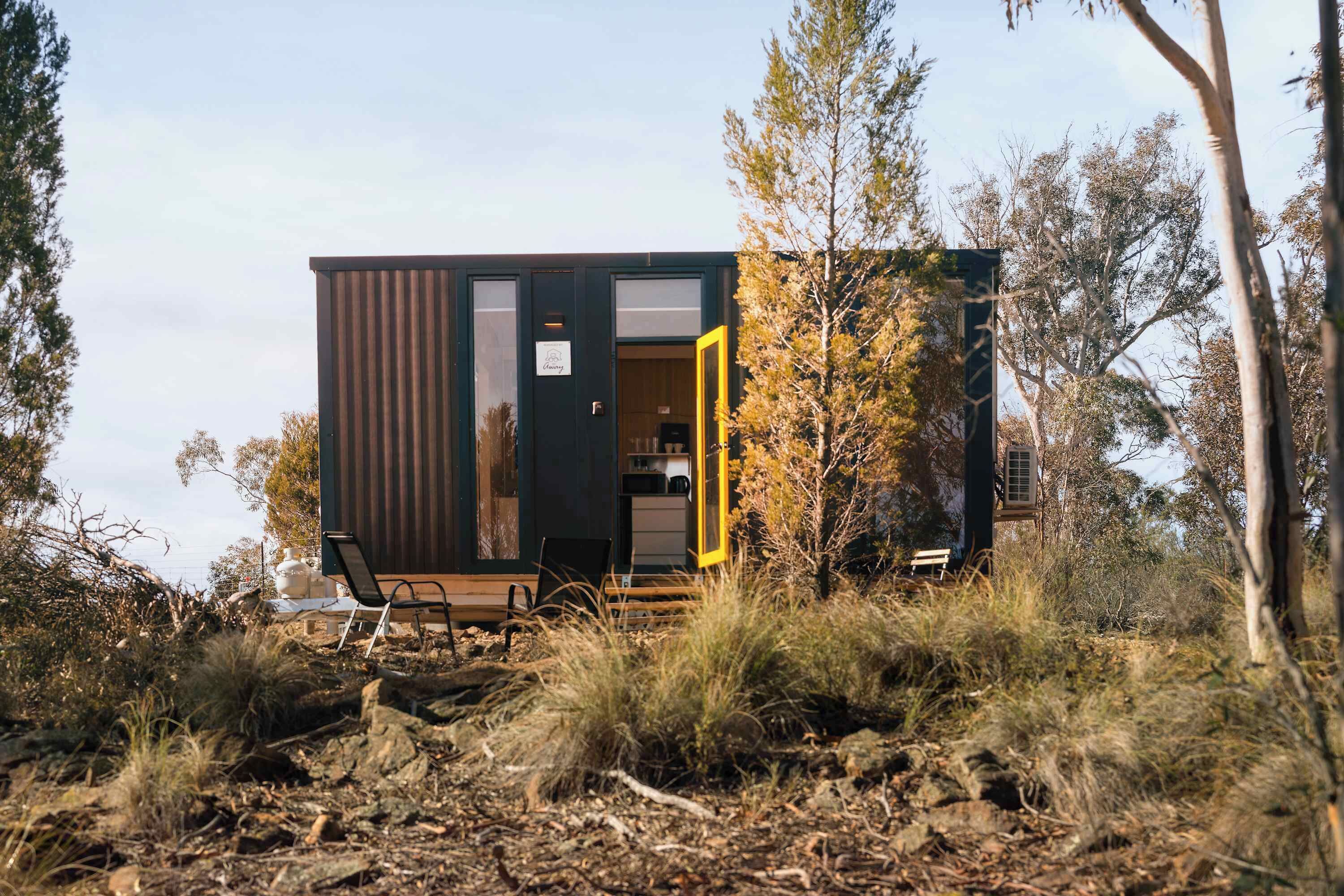 A modern, compact cabin with a large sliding door, situated in a natural setting with trees and shrubs.