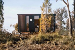 Una cabaña moderna y compacta con un amplio ventanal corredera, ubicada en un entorno natural entre árboles y arbustos.