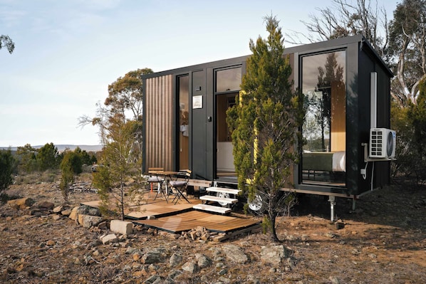 A modern, compact cabin with a large sliding door, a small deck with seating, and an air conditioning unit.