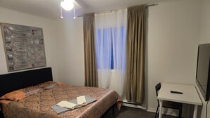 A hotel room with a bed, a desk, a chair, a ceiling fan, a window with curtains, and a wall art piece. - brossard home inn 1 (Brossard)