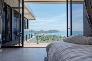 Un dormitorio con una cama grande, un ventanal de vidrio que da a un balcón con vista al mar y las colinas.