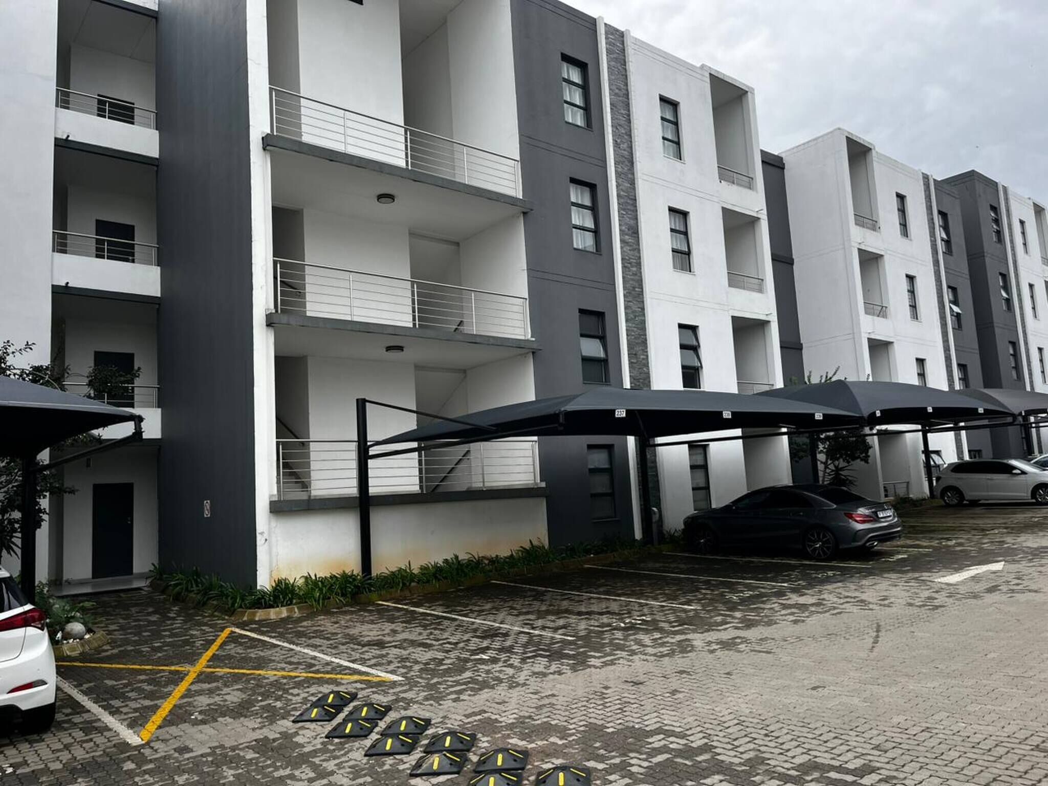 A multi-story residential building with balconies, a parking area with marked spaces, and a covered carport.