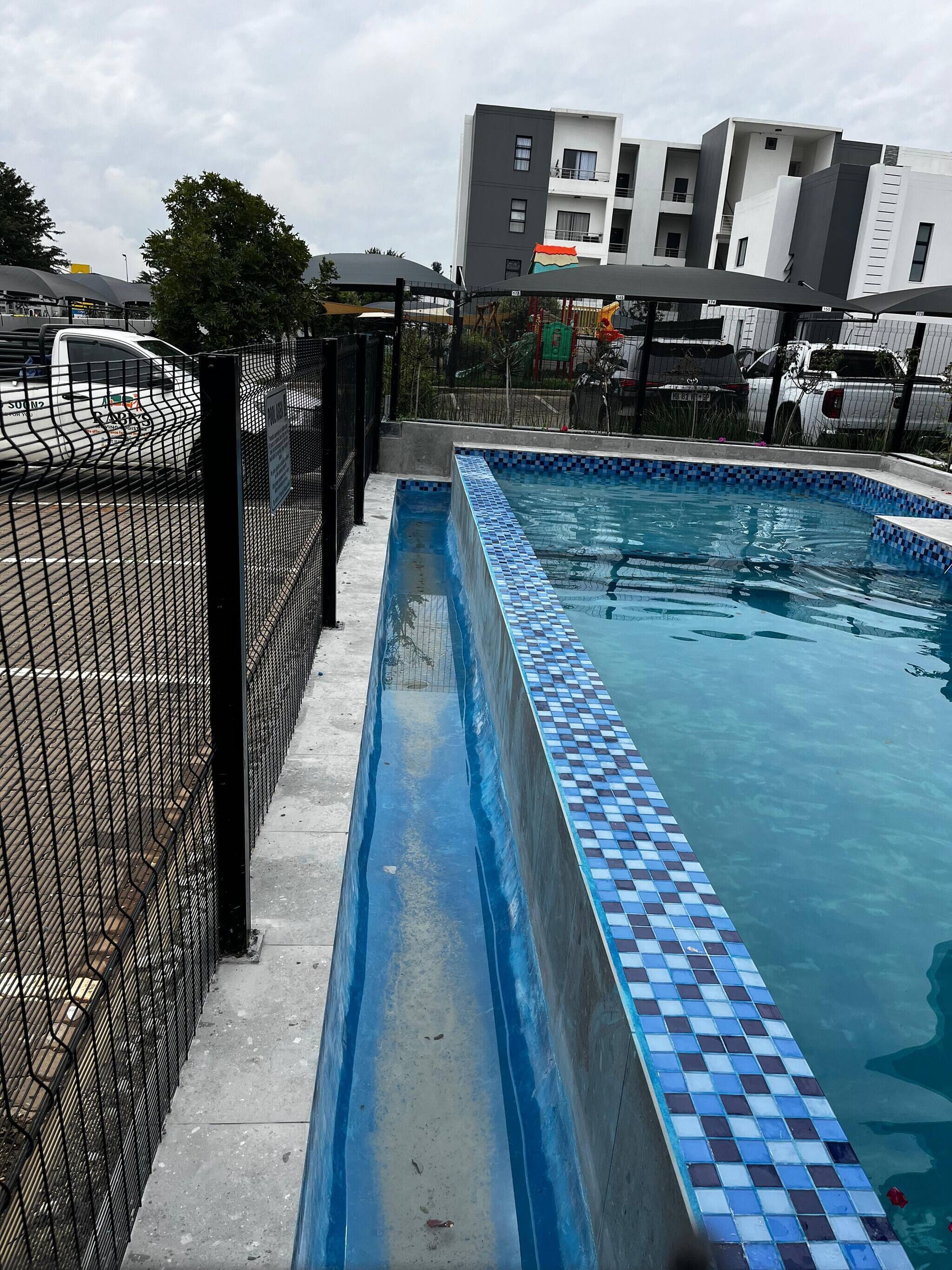 A swimming pool with a tiled edge, surrounded by a black fence, with buildings in the background.
