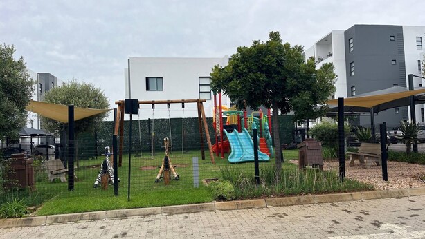 A playground with swings and slides, surrounded by a fence, with buildings in the background. - Stylish 1BR Midrand Pool, Wifi & Secure Parking (Johannesburg)