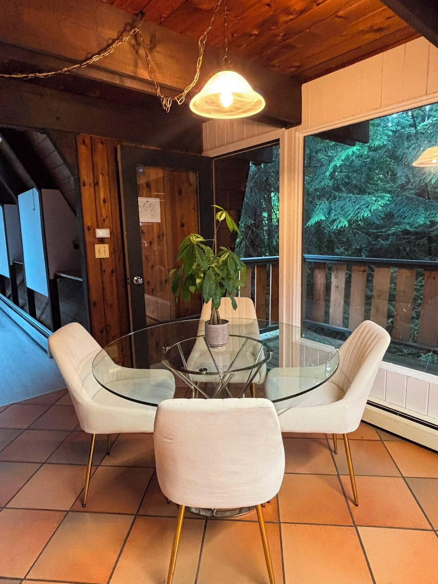 Une pièce avec une table en verre entourée de chaises blanches, une plante en pot et une vue sur une forêt, visible à travers de grandes fenêtres.