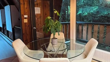 Une pièce avec une table en verre entourée de chaises blanches, une plante en pot et une vue sur une forêt, visible à travers de grandes fenêtres.