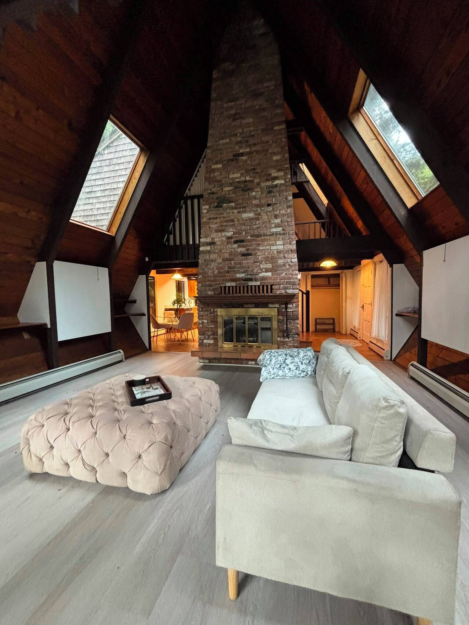 A loft-style room with a stone fireplace, a tufted ottoman, and a white sofa.