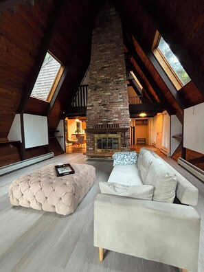 A loft-style room with a stone fireplace, a tufted ottoman, and a white sofa.
