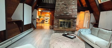 Un loft spacieux avec de hauts plafonds, des poutres apparentes, une cheminée en pierre et une vue sur la cuisine grâce à une porte ouverte.