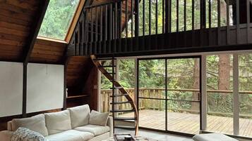 A loft-style room with a high ceiling, a wooden staircase, a sofa, an ottoman, and large windows offering a view of the outdoors.