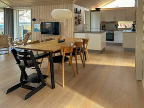 Eine moderne Küche mit Holzschränken, ein Essbereich mit Holztisch und -stühlen, ein an der Wand montierter Fernseher und eine Pendelleuchte.