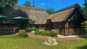 Un bâtiment traditionnel à toit de chaume, doté d’une extension en briques, d’un espace extérieur aménagé pour s’asseoir et d’un chemin en pierre.