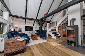 Ein geräumiges Wohnzimmer mit hoher Decke, freiliegenden Balken, einem Kamin und einer Treppe, die zu einer oberen Ebene führt.