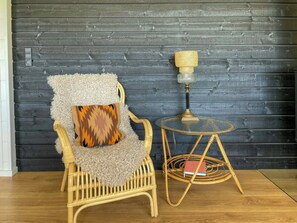 A wicker chair with a fluffy throw blanket and a patterned cushion, a small round table with a glass top and a brass lamp, against a dark wooden wall. - Holiday home with panoramic view of Aarhus Bay (Knebel)