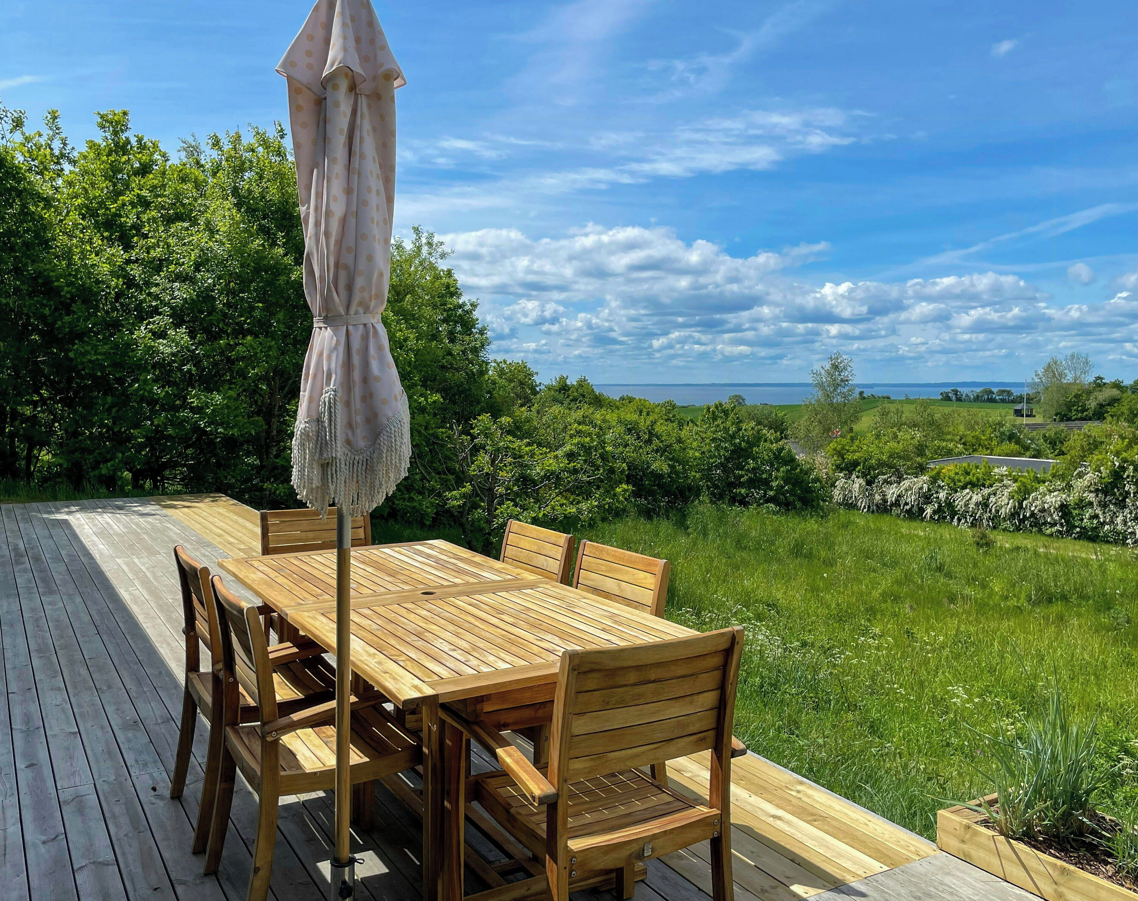 A wooden deck with a table and chairs, an umbrella, and a view of a green landscape and water.