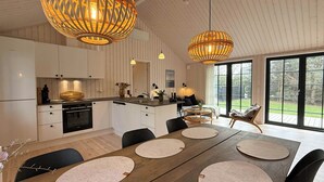 A modern kitchen with white cabinetry, a dining area with a wooden table and chairs, and large windows offering a view of the outdoors. - Modern wooden house built in a classic style (Glesborg)