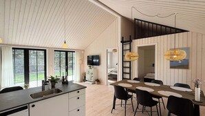 A modern kitchen with a built-in dishwasher, a dining area with a wooden table and black chairs, and a loft bedroom with a black railing. - Modern wooden house built in a classic style (Glesborg)