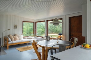 A room with a bed, a dining table, chairs, a lamp, and a view of greenery through large windows. - Adorable retro home with lovely terrace (Bryrup)