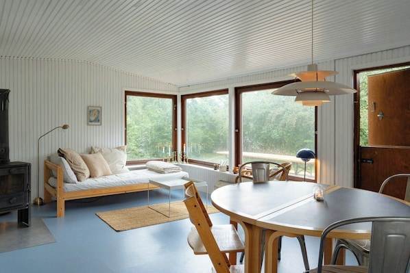 A room with a wooden sofa, a dining table, and a large window with a view of greenery. - Adorable retro home with lovely terrace (Bryrup)