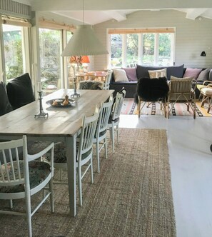 A dining area with a wooden table and white chairs, a sofa with cushions, and a view of greenery outside. - Summerhouse by Tisvilde Hegn forest (Tisvildeleje)