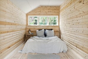 Ein Schlafzimmer mit schrägen Wänden, Holzwänden, einem Bett mit gestreifter Bettwäsche, einem kleinen Nachttisch und einem Fenster mit Blick auf Grünfläche.