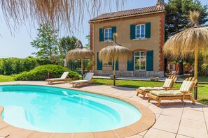 Ein Schwimmbad mit Liegestühlen, ein Haus mit blauen Fensterläden und ein gepflegter Garten.
