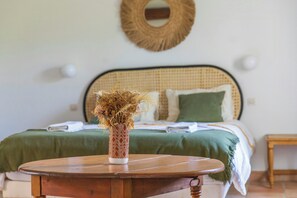 Ein Bett mit grüner Bettdecke, einem Weiden-Kopfstück und einem runden Holztisch mit einer Vase getrockneter Blumen im Vordergrund.