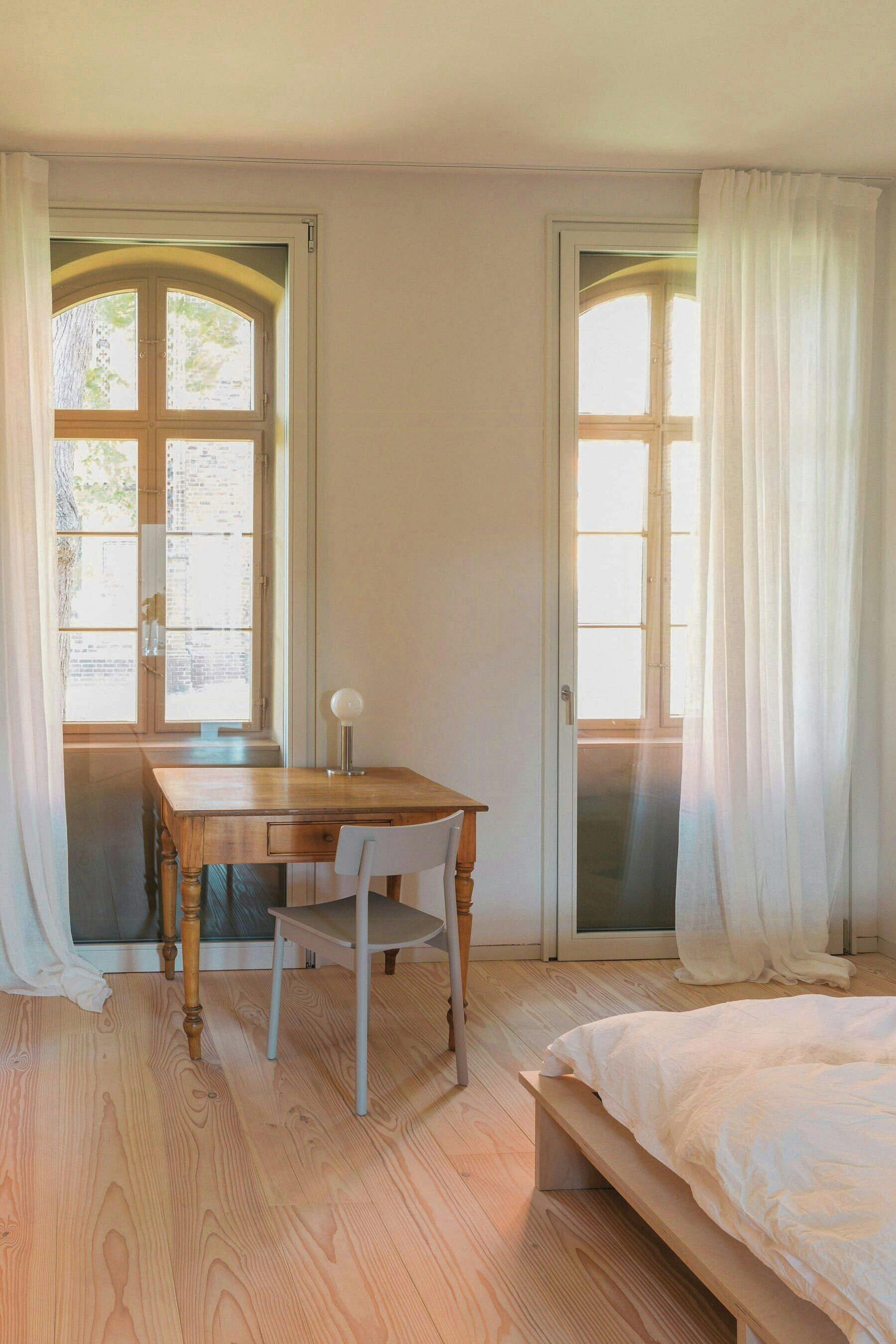 Una habitación con escritorio de madera, una silla, una cama y dos ventanas con cortinas.