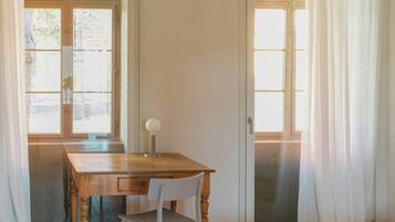 Una habitación con escritorio de madera, una silla, una cama y dos ventanas con cortinas.