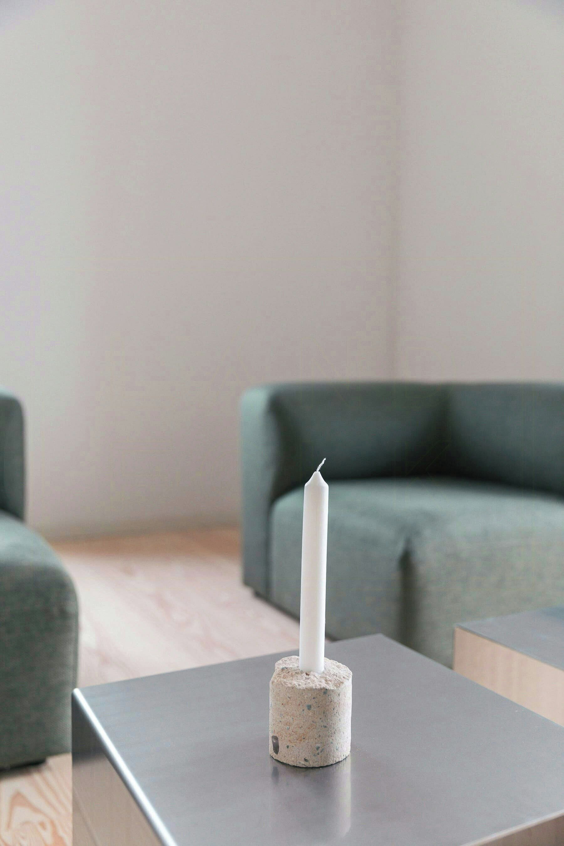 A white candle in a concrete holder on a grey table in a modern room with a green sofa.