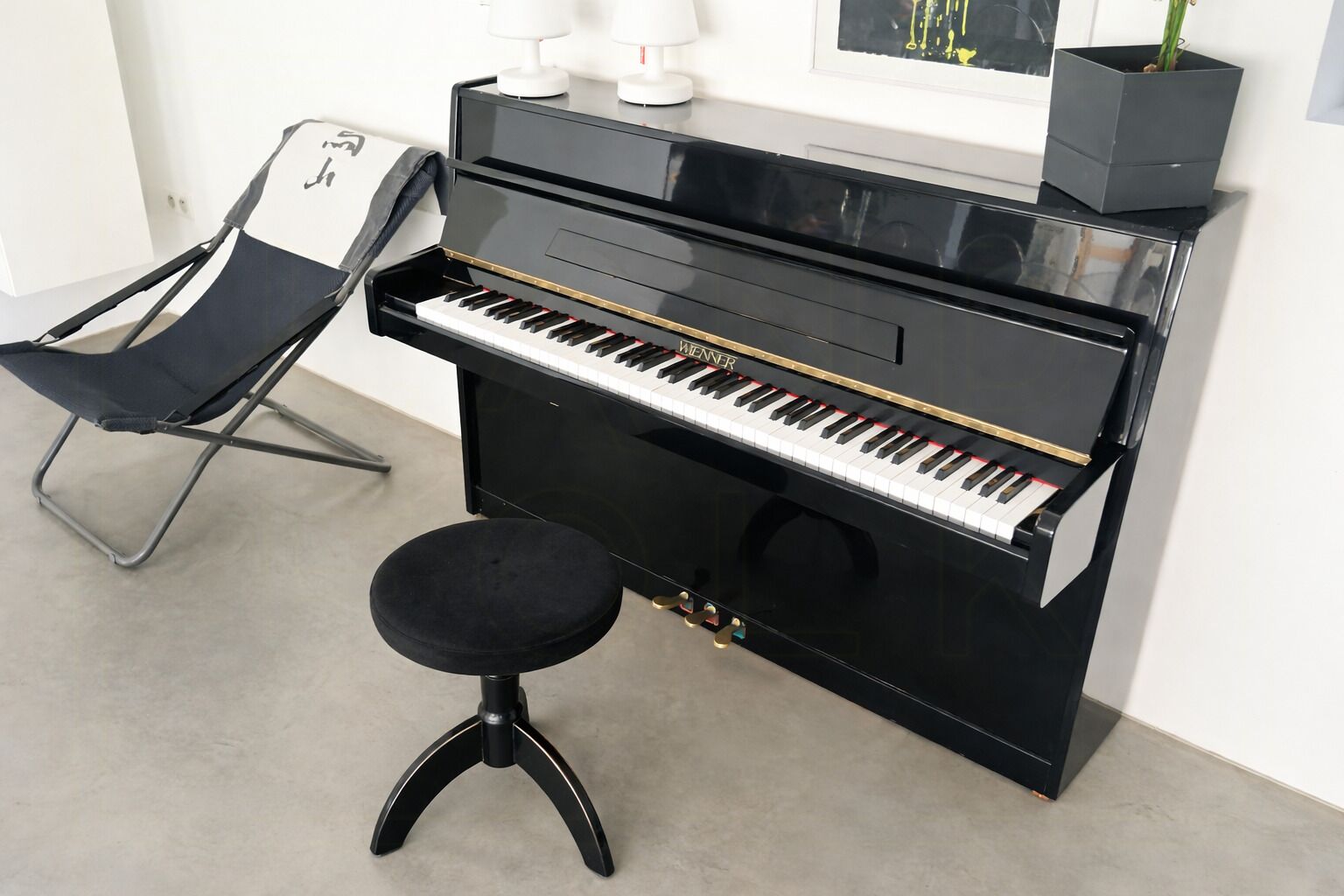 A piano with a stool, a chair, and a potted plant in a room with a white wall and a window.