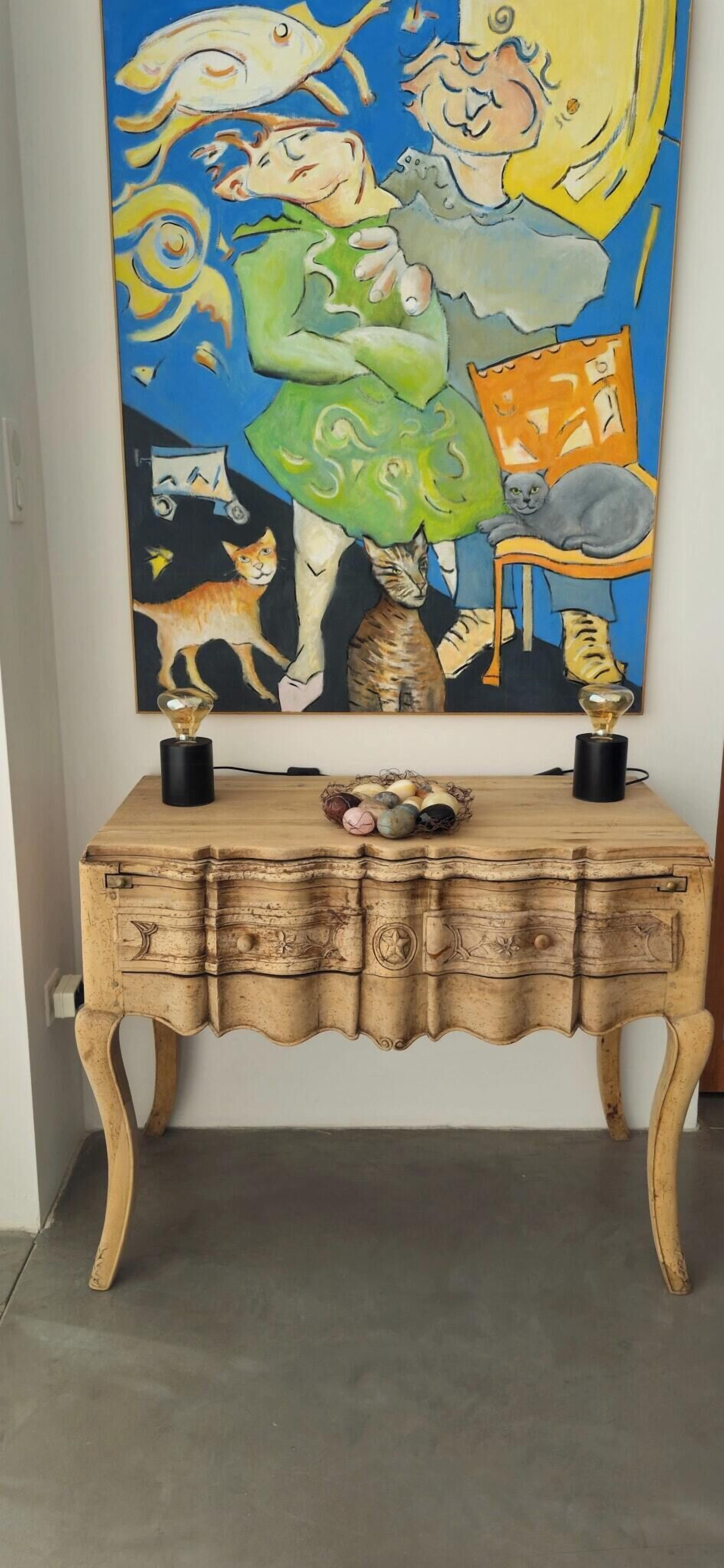 Una consola de madera con dos candelabros negros, un florero con flores y un cuadro abstracto colorido en la parte superior.