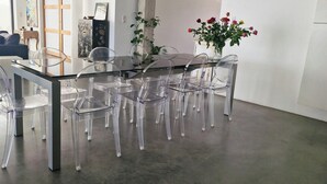 Ein Essbereich mit einem Glastisch, umgeben von transparenten Stühlen, einer Vase mit Blumen und einer Couch im Hintergrund.