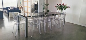 Ein Essbereich mit einem Glastisch, umgeben von transparenten Stühlen, einer Vase mit Blumen und einer Couch im Hintergrund.