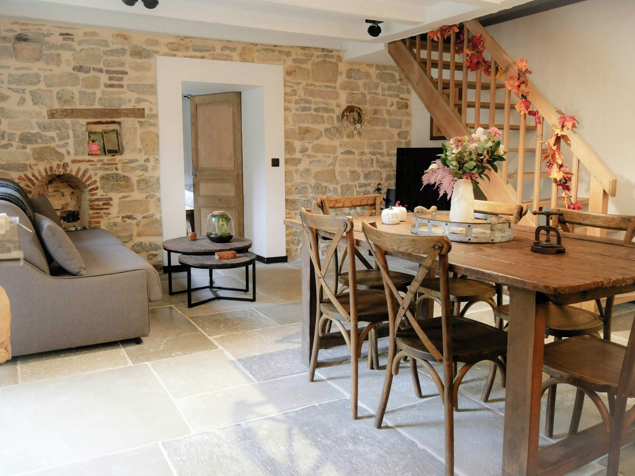 A rustic interior with a stone wall, a wooden dining table, and a staircase with autumn decorations.