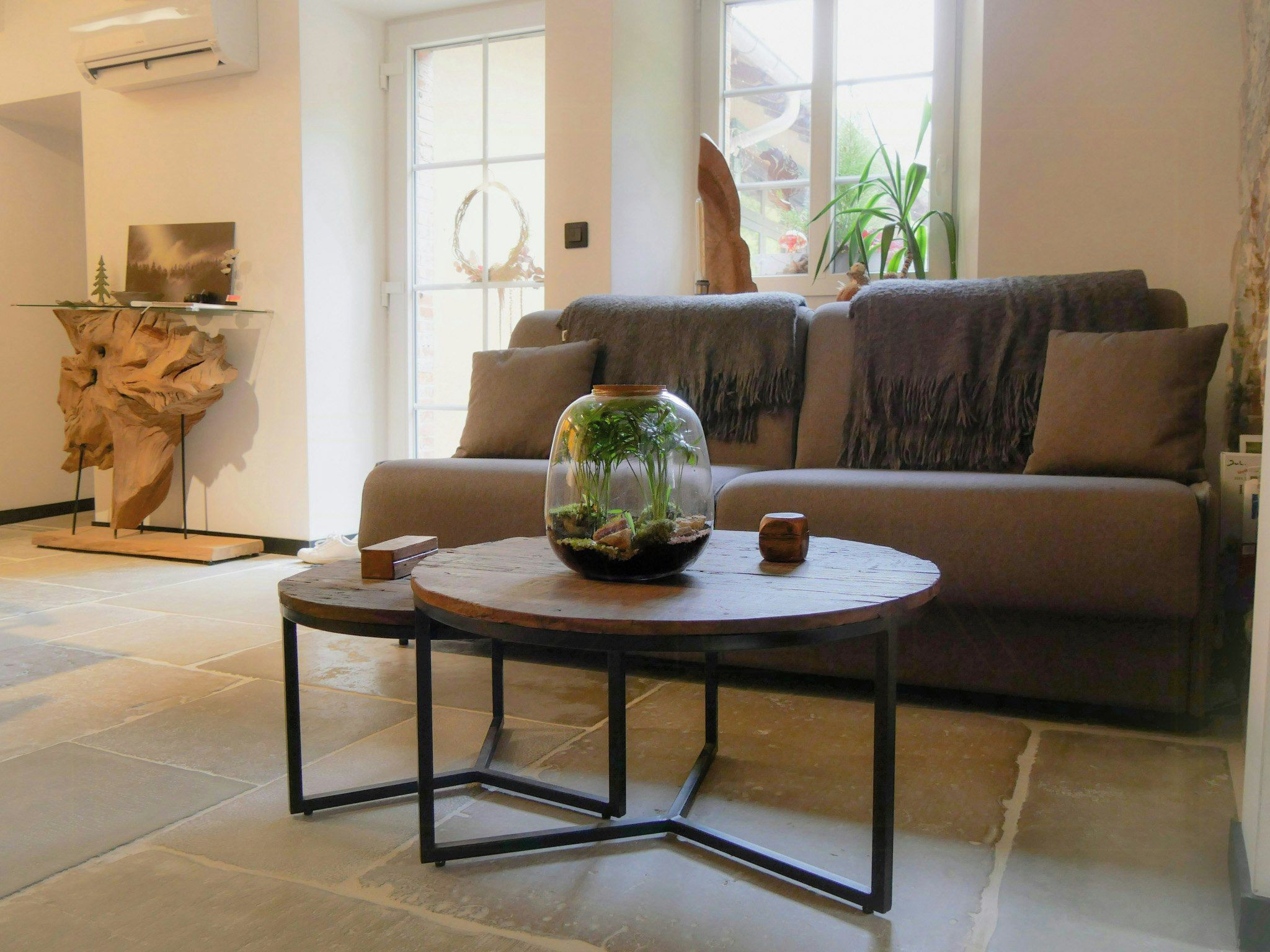 A living room with a sofa, a round table with a glass dome containing plants, and a decorative wooden piece on a stand.
